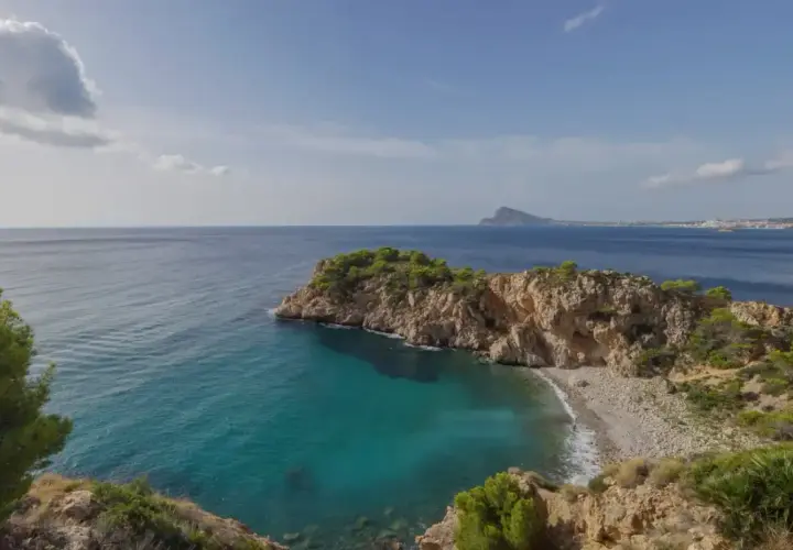 Vista de una cala con aguas turquesas y una gran barra de guijarros, rodeada de acantilados rocosos y vegetación.