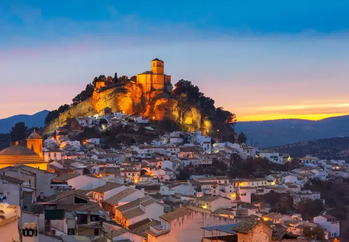 Pueblo blanco andaluz con castillo iluminado en la colina. Un lugar encantador que ver cerca de Granada al atardecer.
