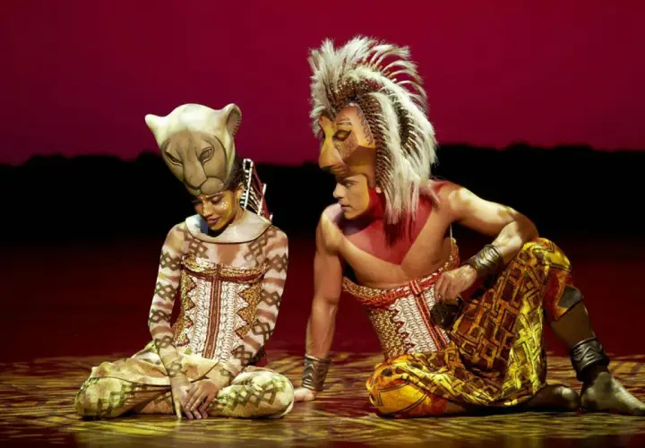 Dos actores con trajes de león, uno con máscara de leona y otro con melena, sentados en el escenario. La duración del Rey León en Madrid es inolvidable.