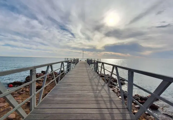 Muelle de madera sobre el mar, con cielo nublado y sol brillante. Un lugar ideal para disfrutar de la playa de Chilches.