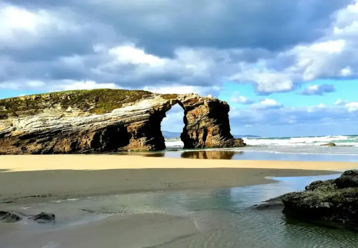 Arco natural en una playa de arena dorada, con el mar y nubes dramáticas. Un ejemplo de las mejores playas Galicia.