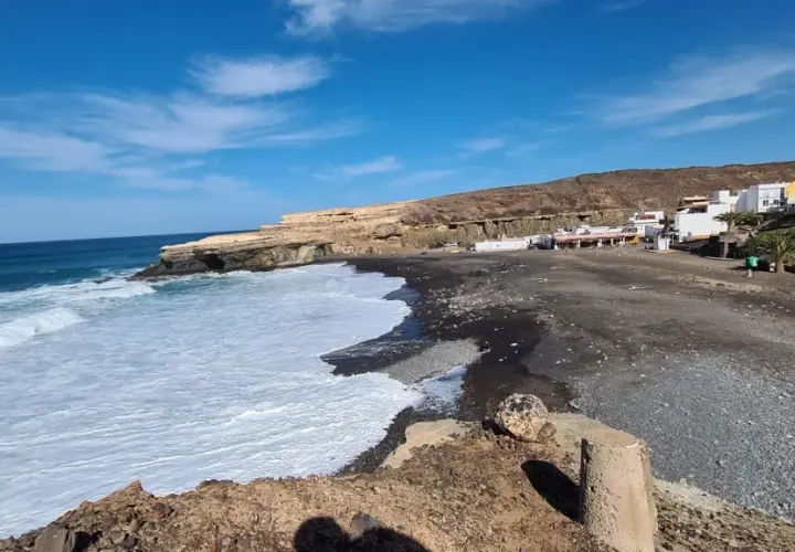 Olas rompen en la playa de arena negra de los charcones de Ajuy, con casas blancas al fondo y acantilados dorados.
