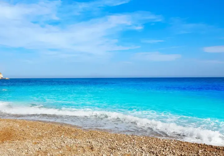 Aguas turquesas bañan la playa de guijarros de cala dels testos, con un acantilado rocoso a la izquierda y un cielo azul despejado.
