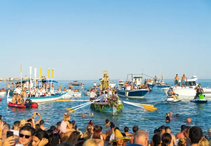 Procesión marítima con la Virgen del Carmen en un barco, rodeada de embarcaciones y gente en el agua, celebrando las fiestas.