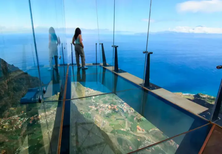 Mujer en mirador de cristal, 3 metros sobre el cielo, con vistas al mar y un pueblo.