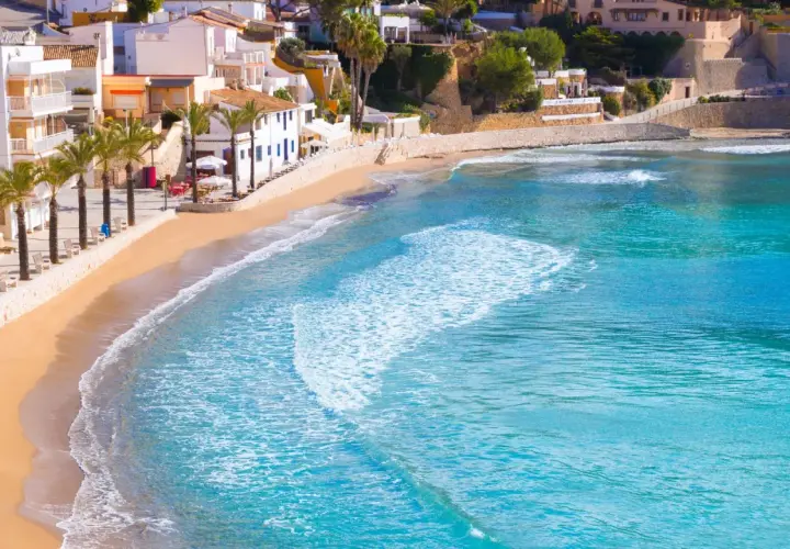 La playa de cala el Portet, con sus aguas turquesas y arena dorada, se extiende bajo un cielo azul, flanqueada por palmeras y casas blancas.