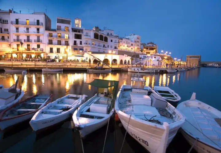 Noche en el paseo marítimo de L'Ametlla de Mar. Barcas blancas flotan en el puerto, reflejando las luces de los edificios y restaurantes.