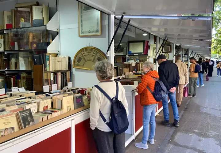 Gente buscando tesoros literarios en la feria del libro antiguo de Madrid. Estanterías repletas de tomos esperan ser descubiertos.