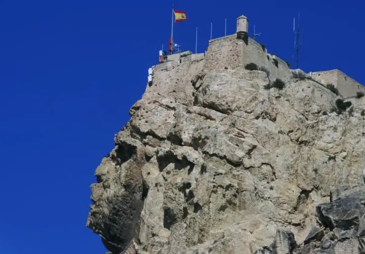 La cara del moro alicante, una roca con forma de perfil humano, corona la cima de una montaña rocosa con un castillo y una bandera española.