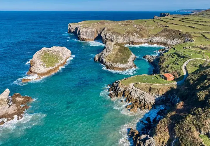 Acantilados y aguas turquesas en la costa de Cantabria. Un lugar idílico que ver en Isla Cantabria, con islotes rocosos y un sendero que lleva a una cabaña.