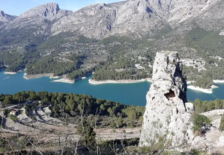 Impresionante vista del embalse de Guadalest con su agua turquesa, rodeado de montañas y pinos. Una roca solitaria se alza como centinela.