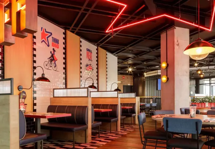 Interior de restaurante con cabinas, mesas y decoración moderna. Las paredes tienen murales inspiradores, como "Think Big". Las luces rojas de neón añaden un toque vibrante. Ideal para leer reseñas de Foster's Hollywood.