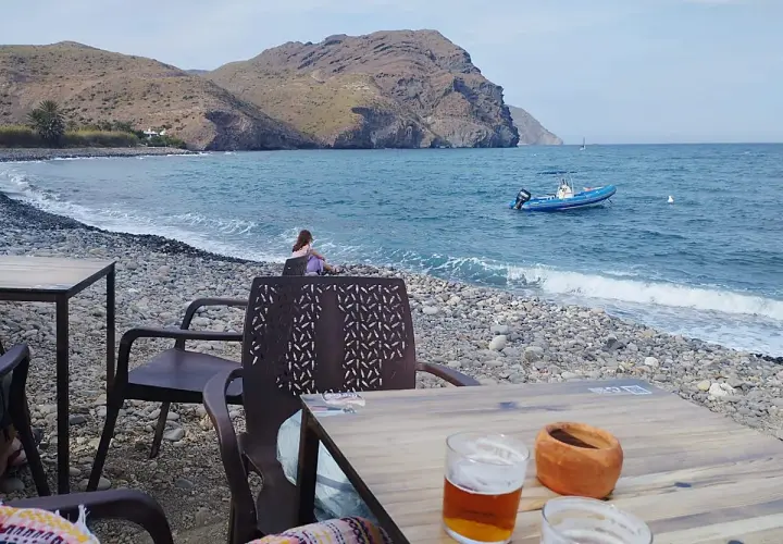 Una persona contempla el mar en la playa de las negras opiniones. Un bote azul descansa en el agua, mientras las olas acarician la costa pedregosa.