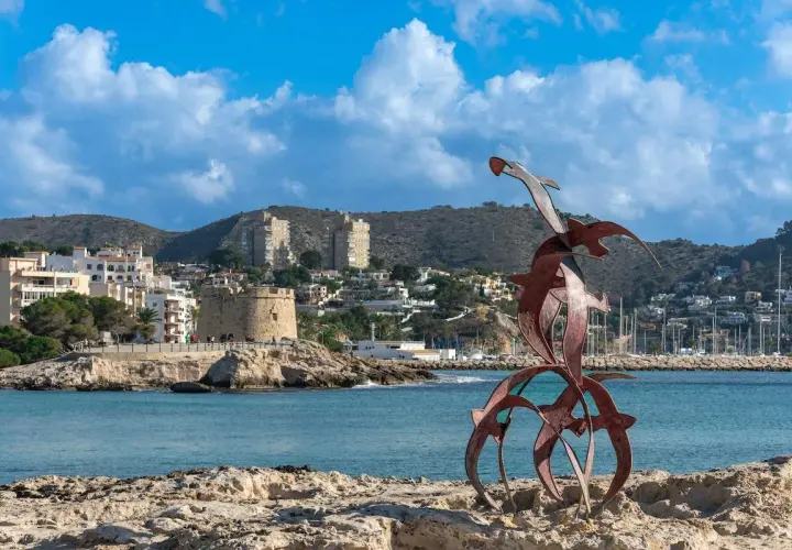 Escultura de aves sobre rocas, con el pueblo de Moraira y su torre defensiva al fondo.