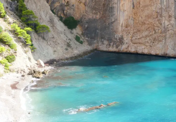 Aguas turquesas bañan la cala racó del corb, rodeada de imponentes acantilados y vegetación verde.
