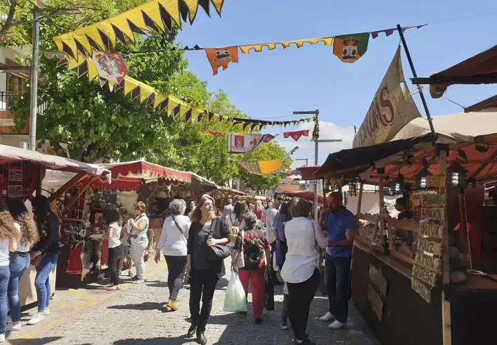 Gente pasea por el mercadillo medieval El Álamo, con puestos de artesanía y comida bajo banderines coloridos.