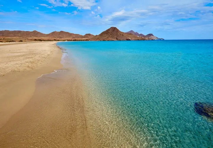 Arena dorada y aguas cristalinas en una de las playas bonitas en España, con montañas áridas al fondo bajo un cielo azul.