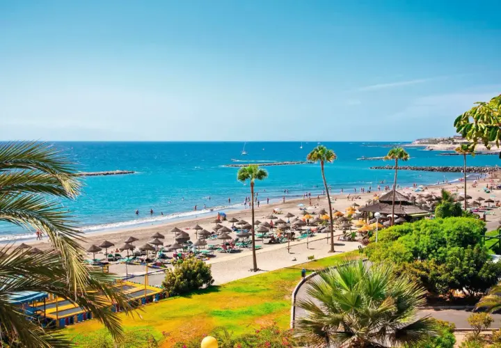 Un día soleado en el paseo marítimo de Playa de las Américas, con gente disfrutando de la playa, sombrillas y palmeras.