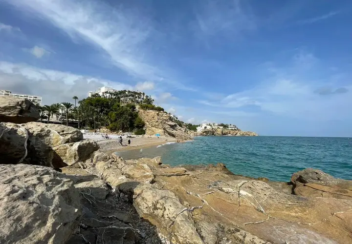 Rocas doradas en primer plano, la caleta Villajoyosa con gente disfrutando del mar azul y edificios blancos en la colina.