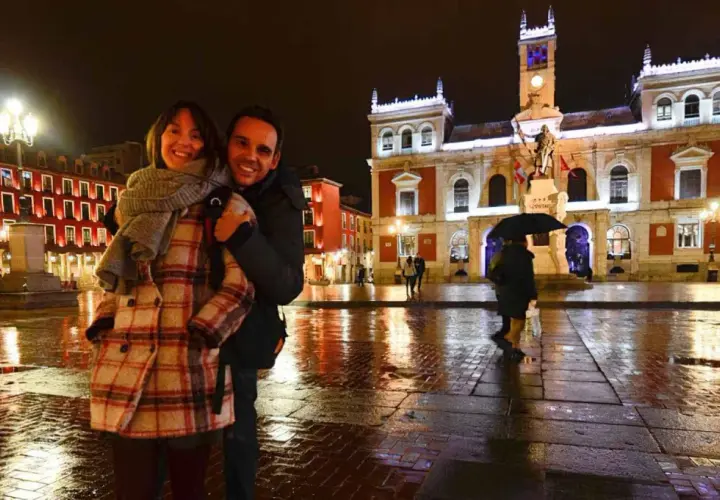 Pareja sonriente en la Plaza Mayor de Valladolid, un lugar bonito que ver en Valladolid, con edificios iluminados y el suelo mojado por la lluvia.