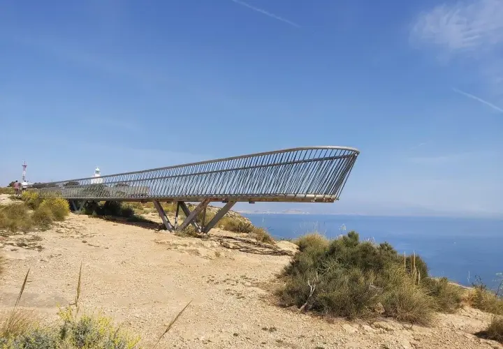 El mirador del faro de Santa Pola se proyecta sobre el mar azul, con vegetación árida y un cielo despejado.