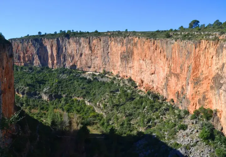 Impresionante pared rocosa anaranjada en la ruta de los calderones Chulilla, con vegetación verde cubriendo el fondo del cañón.