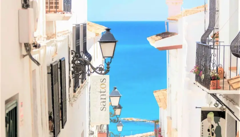 Calle empedrada con vistas al mar azul. Farolas y balcones floridos invitan a descubrir las cosas que hacer en Altea.