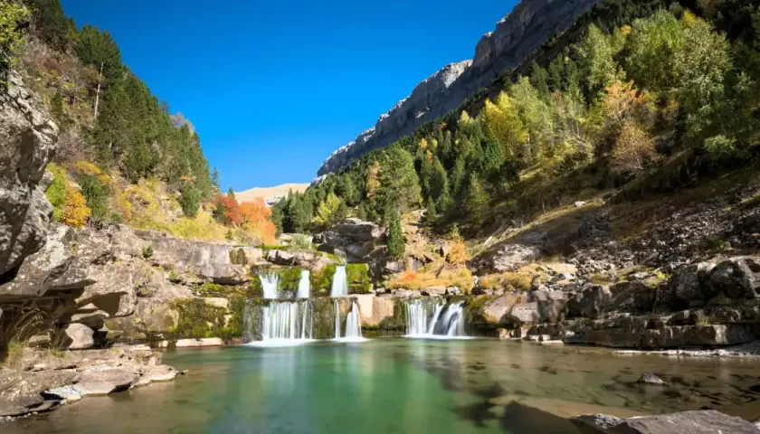 Cascada en un valle montañoso, ideal para rutas senderismo España. El agua verde turquesa fluye entre rocas y árboles otoñales.