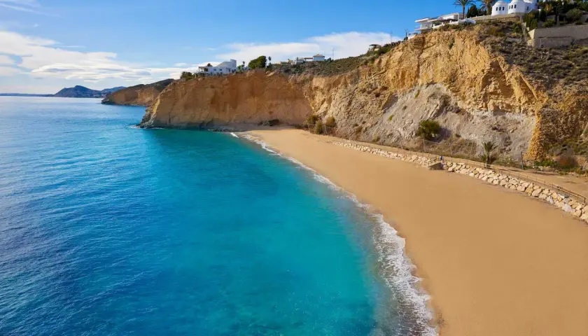 Arena dorada y aguas turquesas en la playa de Villajoyosa, con acantilados y casas blancas al fondo.