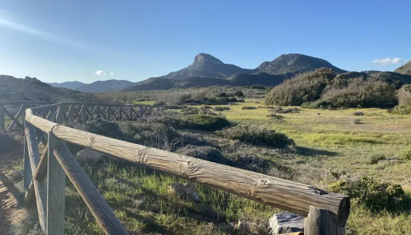Un sendero de madera bordea un paisaje verde y montañoso, con el imponente monte de las cenizas al fondo bajo un cielo azul.