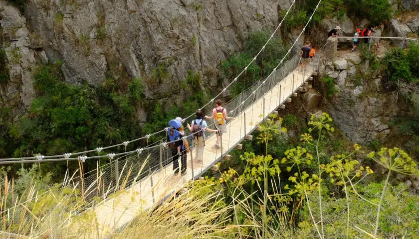 Senderismo en las **rutas Comunidad Valenciana**. Personas cruzan un puente colgante entre riscos y vegetación.