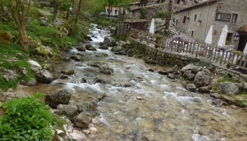 Río caudaloso y rocoso fluye hacia un pueblo de piedra con un cartel que dice "Bar Bulnes Comidas". ¡Así se llega a Bulnes!