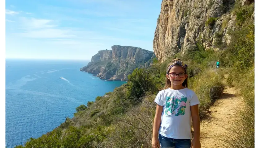 Niña sonriente en la ruta acantilados Benitachell, con el mar azul y formaciones rocosas al fondo.