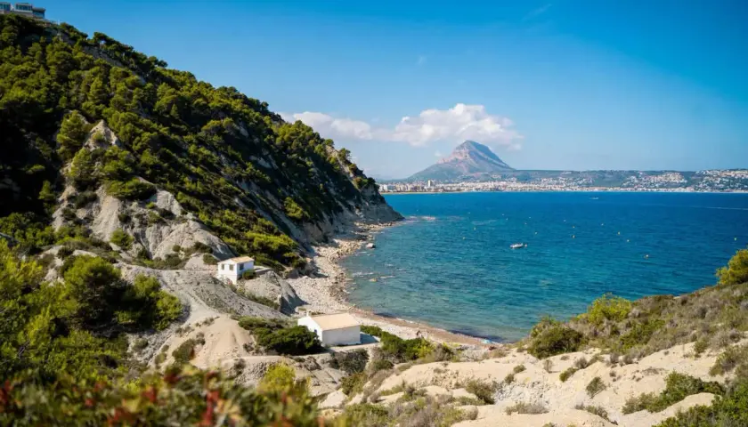 La cala del francés, con sus casitas blancas y el mar azul, se asoma a un paisaje rocoso y verde. Al fondo, la silueta de una montaña domina la costa.