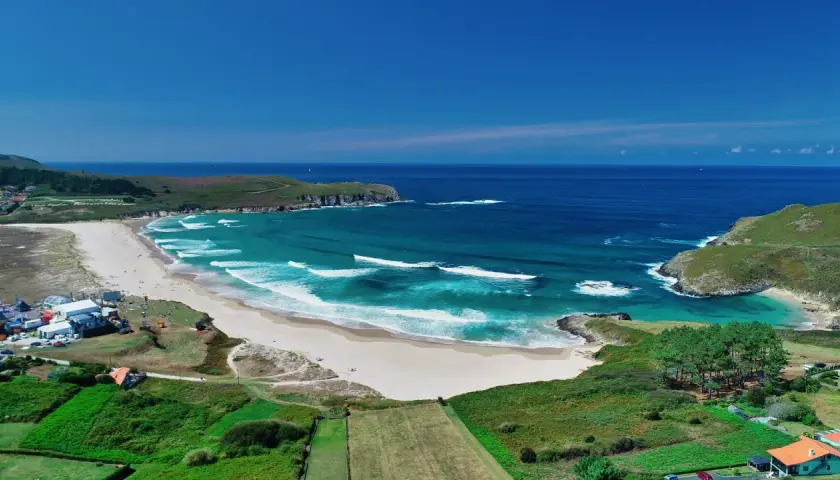 Vista aérea de la playa de Pantín con olas turquesas rompiendo en la arena dorada, rodeada de verdes colinas y el azul intenso del océano.