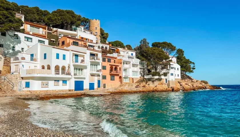 Casas blancas y coloridas se asoman al mar turquesa en la costa brava españa. Una pequeña playa de guijarros recibe las olas.