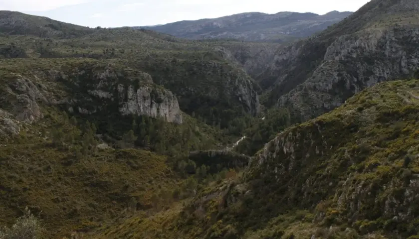 Paisaje montañoso con vegetación verde y rocas escarpadas. El **barranc de l'infern** se abre paso entre las colinas, con un camino serpenteante.