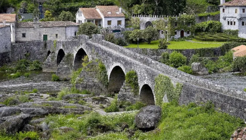Puente de piedra cubierto de hiedra, con casas tradicionales al fondo. Un lugar idílico para visitar, así es como ponte Maceira como llegar.