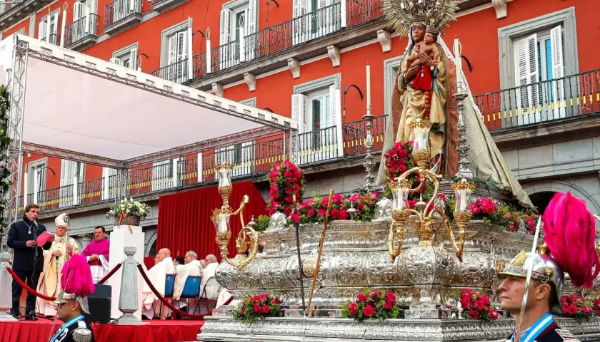 Procesión de la Almudena en Madrid. Una imagen de la Virgen con el Niño sobre un trono adornado con flores, con el fondo de edificios rojos y gente vestida de gala.