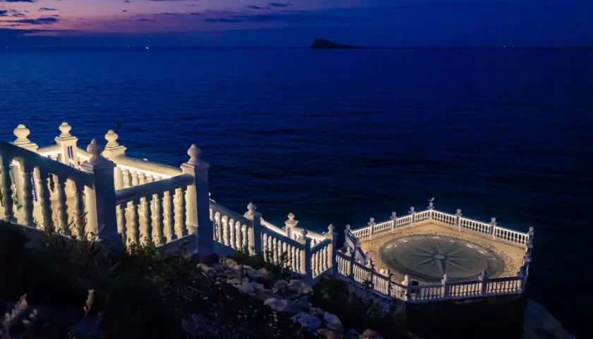 El mirador de Benidorm de noche, con el mar azul oscuro y un cielo crepuscular. Una balaustrada blanca ilumina el camino hacia una plataforma circular con una rosa de los vientos.