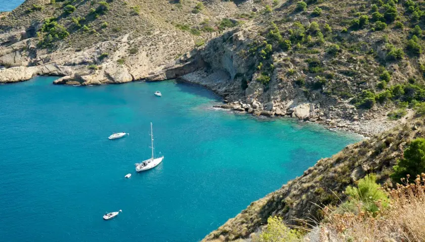 La cala del metge, con aguas turquesas y barcos navegando, se abre paso entre acantilados rocosos y vegetación mediterránea.
