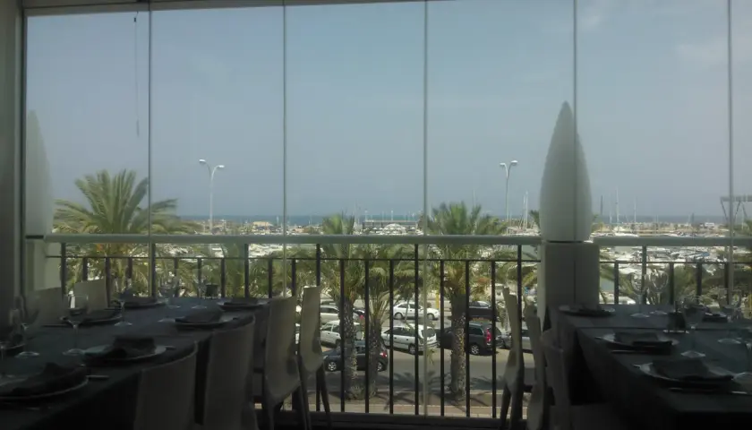 Mesas puestas con vistas al puerto deportivo de la barqueta Denia. Palmeras y barcos crean un ambiente vacacional.