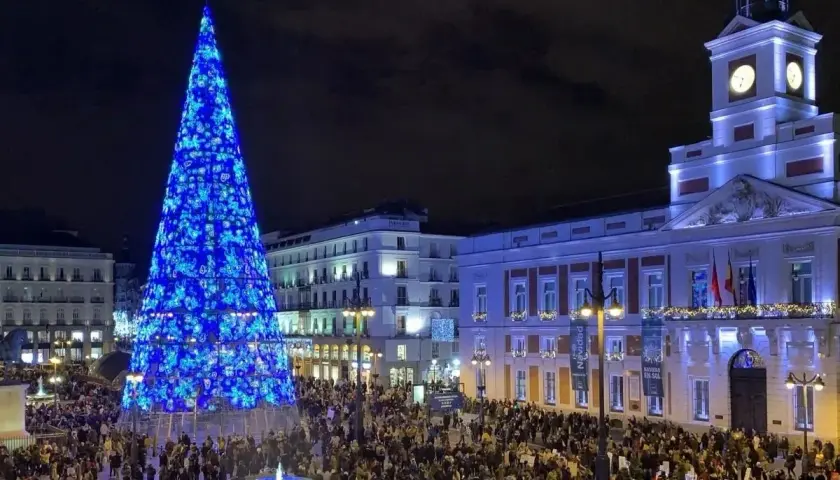 Gran árbol de Navidad azul ilumina la Puerta del Sol, llena de gente, ideal para planes de Navidad en Madrid.