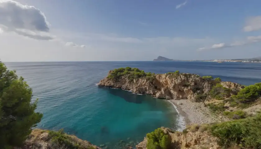 Vista de una cala con aguas turquesas y una gran barra de guijarros, rodeada de acantilados rocosos y vegetación.