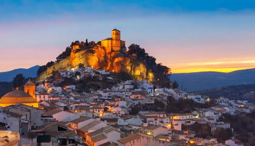 Pueblo blanco andaluz con castillo iluminado en la colina. Un lugar encantador que ver cerca de Granada al atardecer.