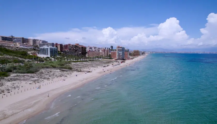 Vista aérea de la playa los Arenales con edificios residenciales al fondo y el mar turquesa.