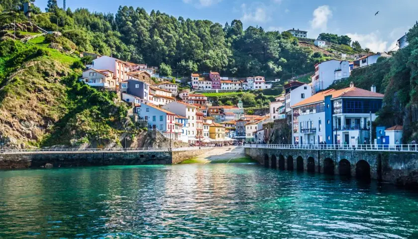 Pueblo costero con casas de colores en una ladera verde, un puerto y aguas turquesas. Un ejemplo de los lugares más bonitos de España.