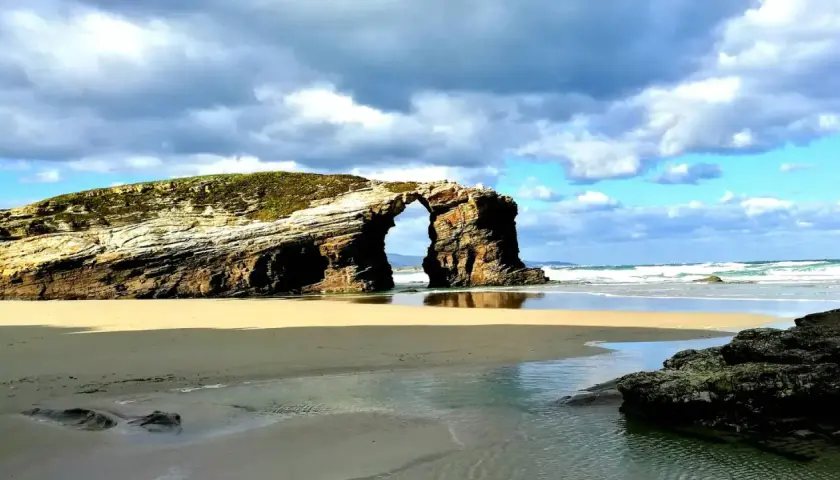 Arco natural en una playa de arena dorada, con el mar y nubes dramáticas. Un ejemplo de las mejores playas Galicia.