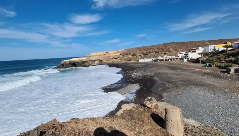 Olas rompen en la playa de arena negra de los charcones de Ajuy, con casas blancas al fondo y acantilados dorados.