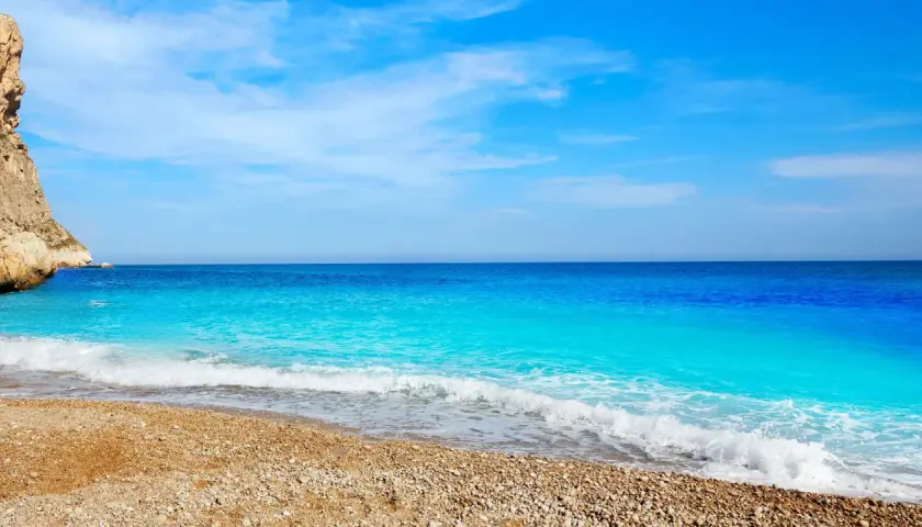 Aguas turquesas bañan la playa de guijarros de cala dels testos, con un acantilado rocoso a la izquierda y un cielo azul despejado.
